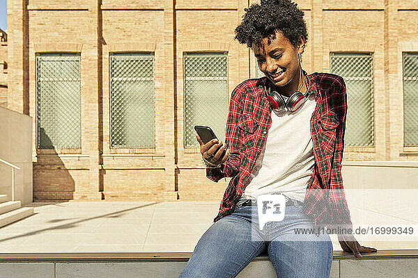 Smiling young woman using mobile phone while sitting against building