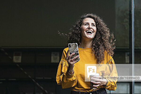 Fröhliche Frau mit lockigem Haar  die lachend ein Mobiltelefon und einen Einwegbecher hält