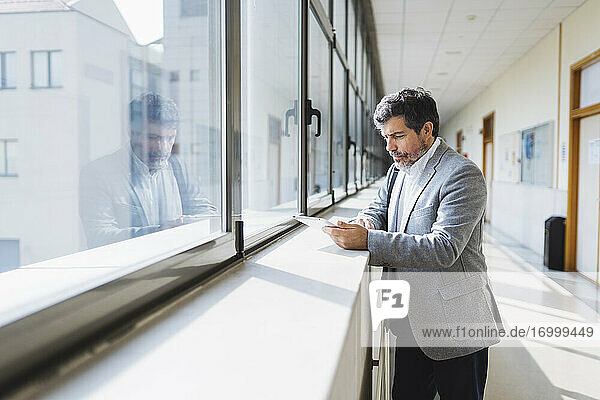 Professor  der ein digitales Tablet benutzt  steht auf dem Fensterbrett im Korridor einer Universität