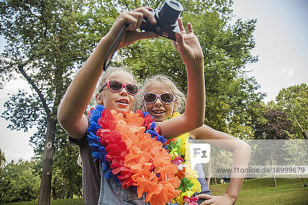 Fröhliche Mädchen mit Sonnenbrillen und Blumengirlanden benutzen die Kamera im Park