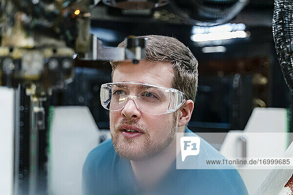 Male worker wearing protective eyewear while examining robotic arm at industry