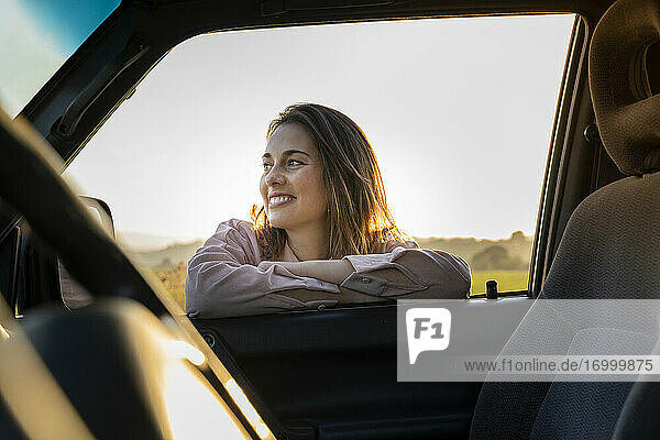 Happy young woman looking away while leaning on car window during sunset