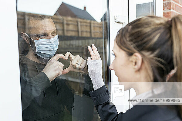 Freund zeigt seiner Freundin durch das Fensterglas eine herzförmige Geste  während er zu Hause unter Quarantäne steht