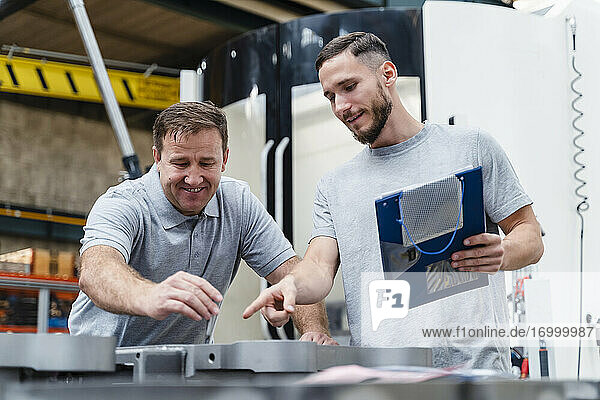 Mature male technician doing quality control of product while standing with young coworker at illuminated factory