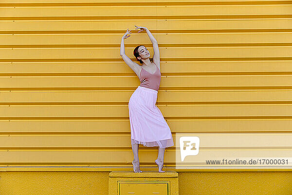 Ballerina mit erhobenen Armen in Pose  während sie auf einem Sitz vor einer gelben Wand steht