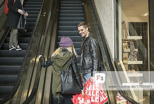 Young couple carrying shopping bags while standing on escalator in shopping mall