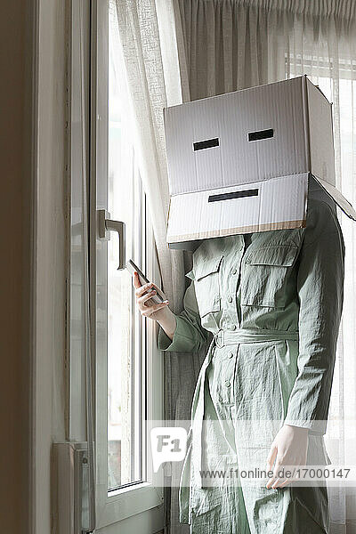 Woman wearing a cardbox on head with bored smiley looking out of the window and using smartphone