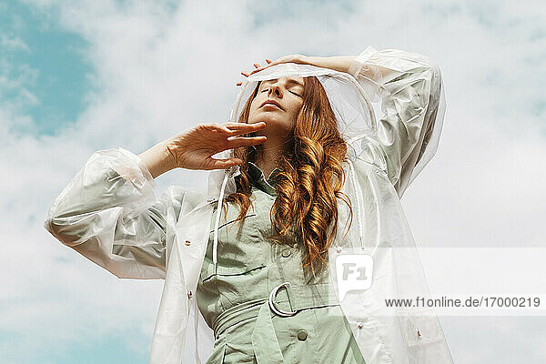 Rothaarige junge Frau mit geschlossenen Augen  die einen durchsichtigen Regenmantel trägt und vor einem bewölkten Himmel steht