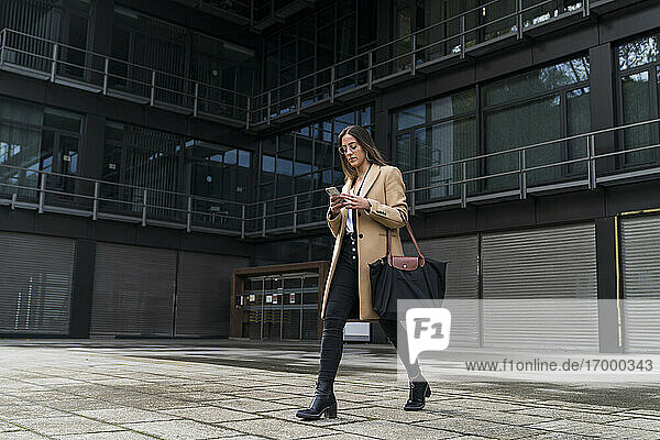Fashionable businesswoman using mobile phone while walking on footpath against office building