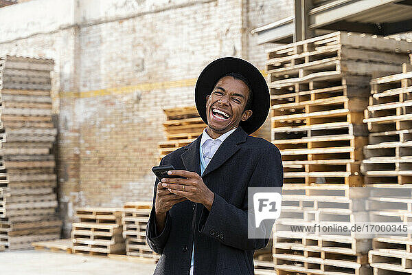 Junger Mann  der lachend sein Handy gegen Holzpaletten hält