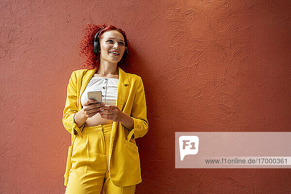 Junge Frau in gelbem Anzug lehnt an einer roten Wand  trägt Kopfhörer und benutzt ein Smartphone
