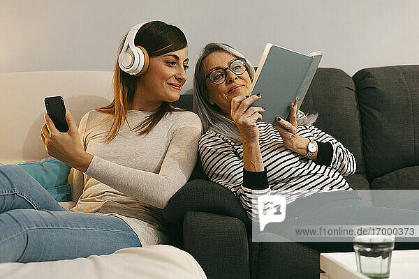 Mutter zeigt ihrer Tochter ein Buch und benutzt dabei ihr Handy  während sie zu Hause auf dem Sofa sitzt