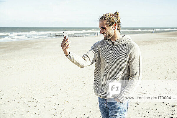 Junger Mann lächelt  während er ein Selfie mit seinem Handy am Strand macht
