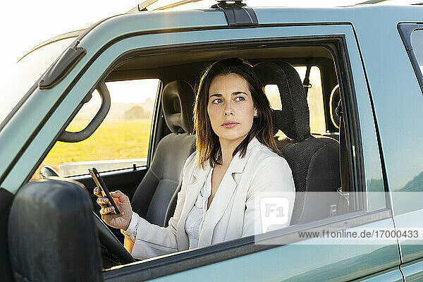 Junge Frau  die durch das Fenster schaut  während sie ihr Handy im Auto während einer Autofahrt hält