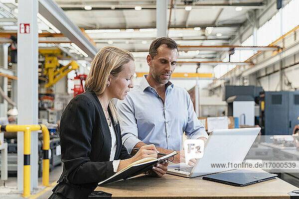 Unternehmerin mit Buch  während ein männlicher Kollege in einer Fabrik am Laptop arbeitet