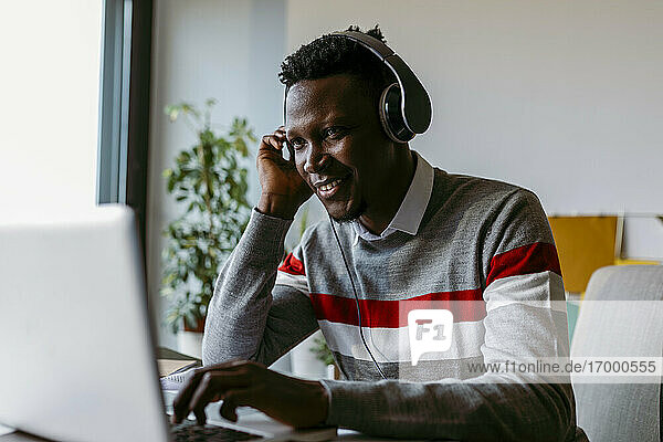 Lächelnder männlicher Unternehmer mit Kopfhörern  der zu Hause einen Laptop benutzt
