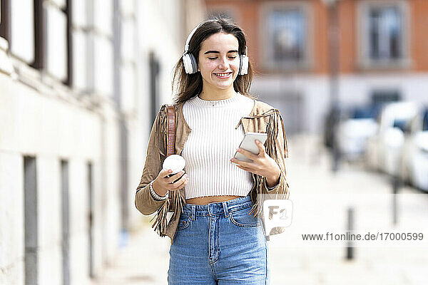 Lächelnde Frau mit Kopfhörern und Smartphone auf dem Fußweg