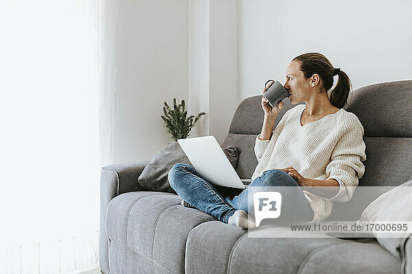 Frau  die wegschaut und Kaffee trinkt  während sie mit ihrem Laptop zu Hause auf dem Sofa sitzt