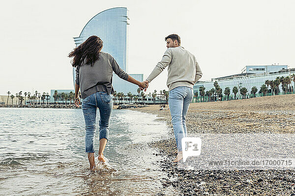 Freundin und Freund halten sich an den Händen  während sie am Strand im Wasser spazieren gehen
