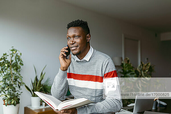 Businessman talking on smart phone while holding book at home office