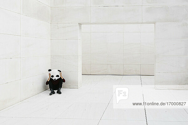 Frau mit Panda-Maske sitzt in einer Ecke vor einer weißen Wand