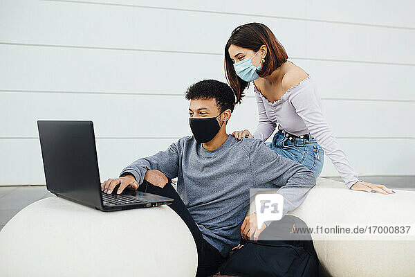 Young couple wearing face mask using laptop while sitting by white concrete ball against wall