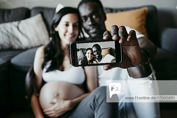 Lächelnder junger Mann mit schwangerer Frau macht Selfie zu Hause