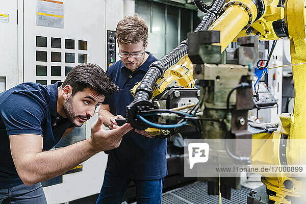 Male coworkers examining robotic arm while standing in manufacturing factory