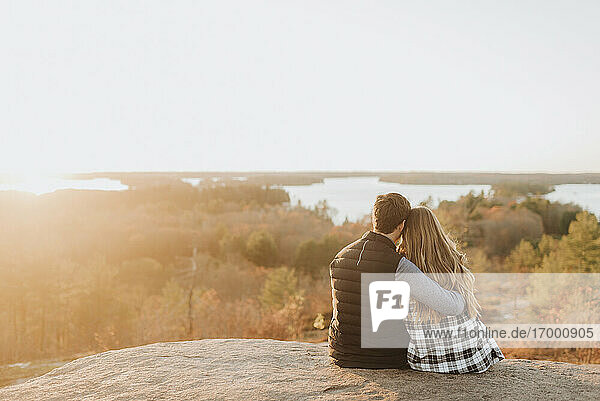 Junges Paar sitzt zusammen auf einem Felsen und bewundert die untergehende Sonne im Herbst
