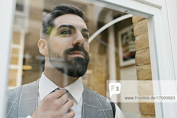 Young groom adjusting necktie while looking through window at home