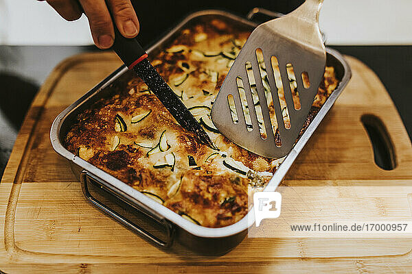 Nahaufnahme einer Frau  die gekochte Lasagne mit Zucchini in einem Tablett zu Hause von Hand schneidet