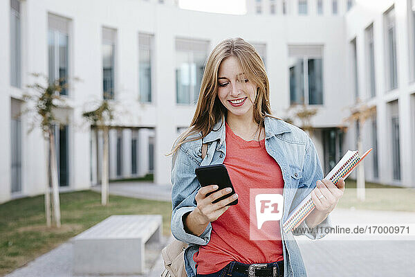 Smiling young blond female student using smart phone at university