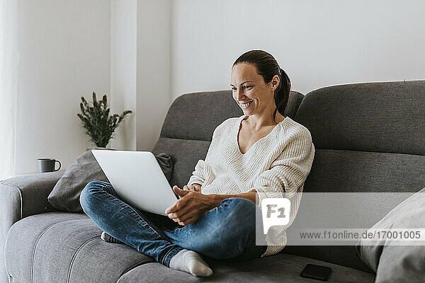 Smiling woman sitting cross-legged using laptop on sofa in living room