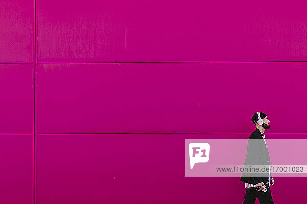 Young man with headphones and smartphone walking along a pink wall