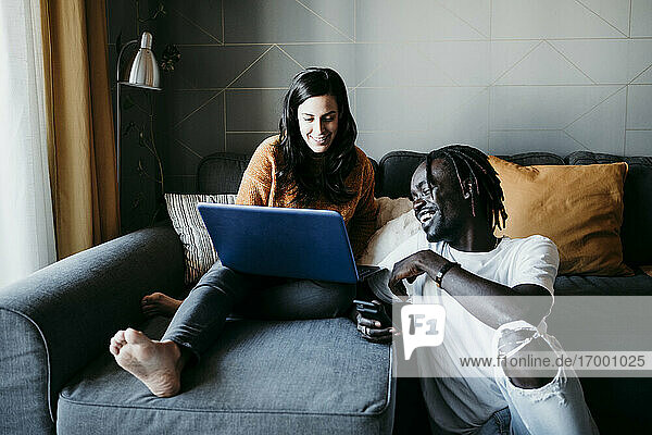 Smiling young man with pregnant woman using laptop while sitting on sofa at home