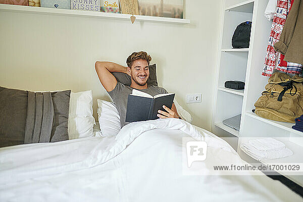Lächelnder Mann mit Hand hinter dem Kopf  der im Schlafzimmer zu Hause ein Buch liest