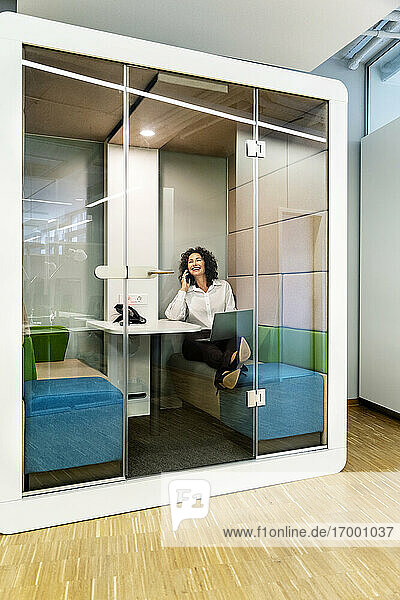 Geschäftsfrau mit Laptop  die in einer Kabine im Büro sitzt und mit ihrem Mobiltelefon spricht