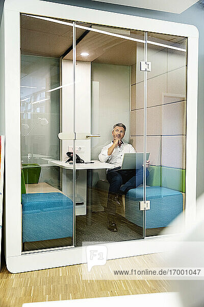 Nachdenklicher Geschäftsmann mit Laptop  während er in einer Kabine im Büro sitzt