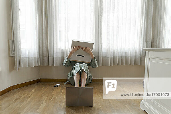 Woman wearing a cardbox on head with bored smiley sitting on floor in front of laptop  covering the eyes