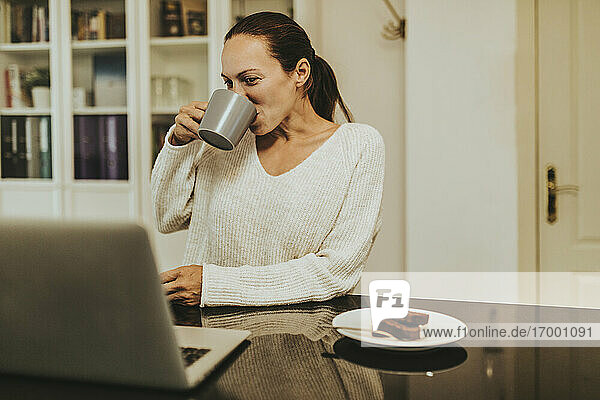 Frau trinkt Kaffee und schaut auf einen Laptop in einer beleuchteten Küche