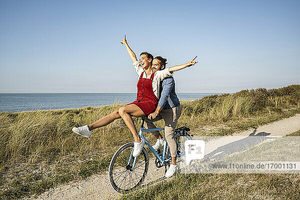 Frau mit erhobener Hand auf dem Lenker sitzend  während sie eine Fahrradtour mit ihrem Freund gegen den klaren Himmel genießt