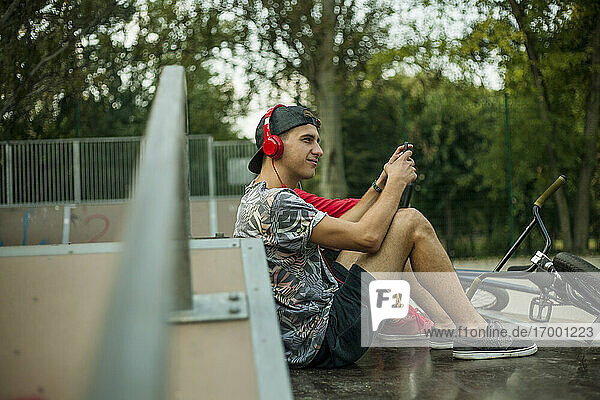 Lächelnder junger Mann hört Musik  während er mit einem Freund im Skateboardpark sitzt