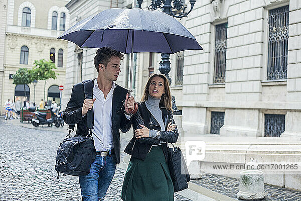 Geschäftsfrau mit männlichem Kollegen  die unter einem Regenschirm auf einer Straße in der Stadt spazieren gehen