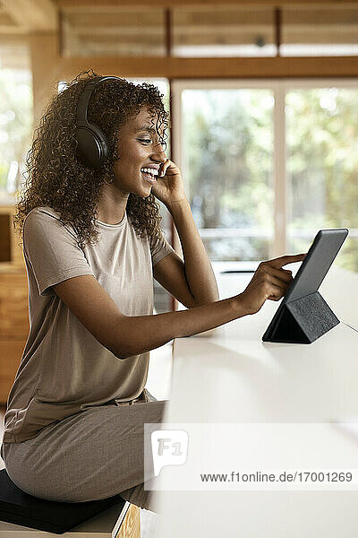 Lächelnde Geschäftsfrau mit digitalem Tablet  die über Kopfhörer spricht  während sie zu Hause sitzt