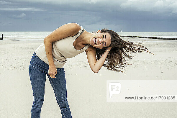 Fröhliche Frau mit Hand im Haar am Strand stehend