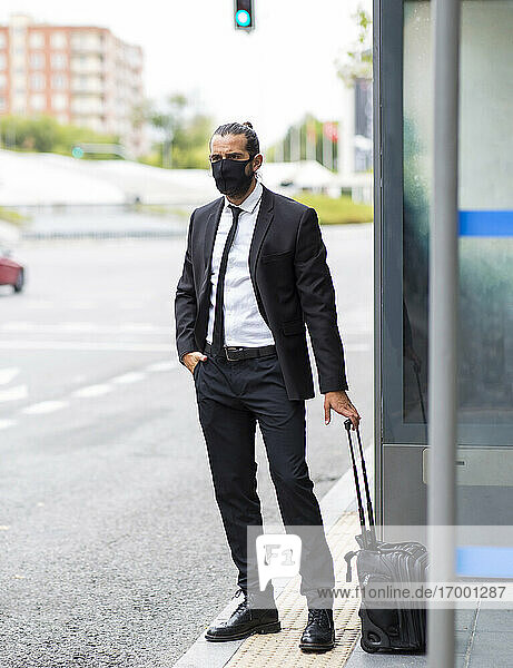 Geschäftsmann mit Gesichtsschutzmaske  der mit einem Koffer an der Bushaltestelle wartet