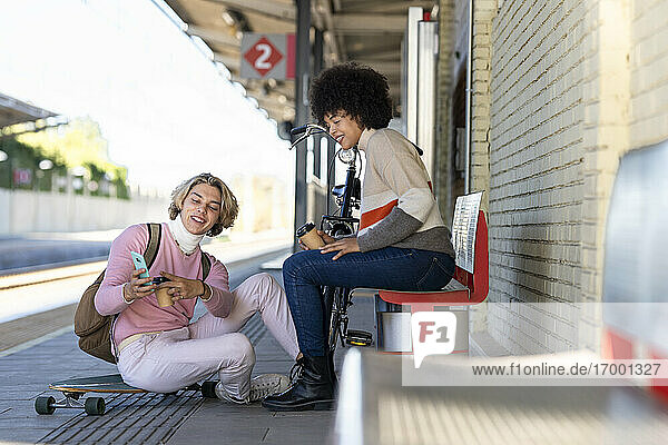 Junger Mann teilt sein Smartphone mit einer Freundin  während er am Bahnhof sitzt