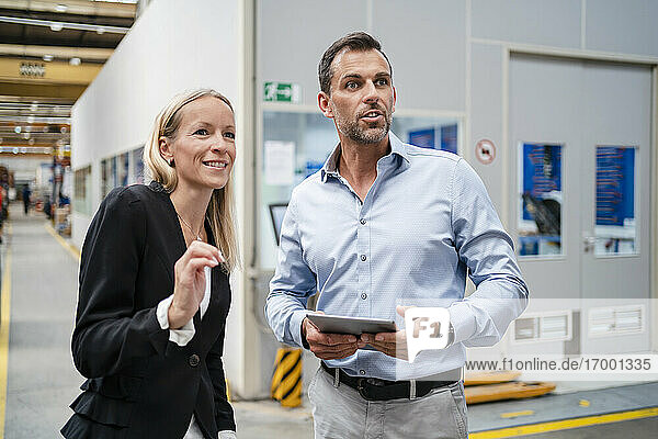 Smiling female entrepreneur and businessman looking away while holding digital tablet standing at industry