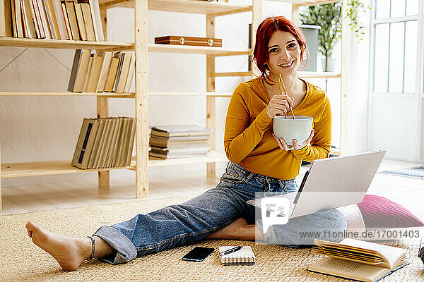 Lächelnde Frau beim Essen und Lernen am Laptop zu Hause sitzend