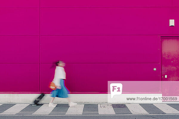 Junge Frau mit Smartphone geht mit Trolley entlang einer rosa Wand  unscharf